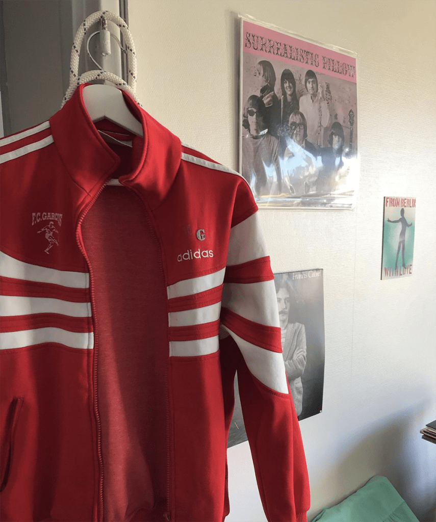 Veste rouge Adidas accrochée dans une chambre, parmi les couvertures de vinyles au mur. Logo du FC Garches 